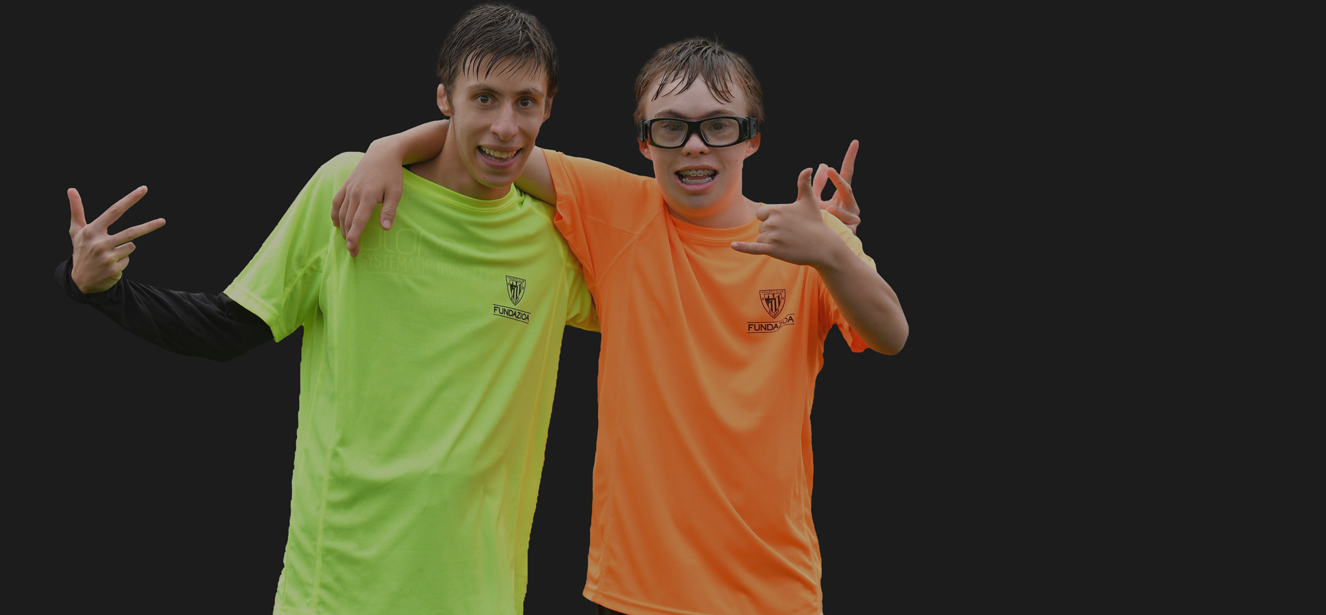 Dos jóvenes con camisetas de la Fundación Athletic Club mostrando señales con las manos, representando inclusión y alegría en actividades deportivas.